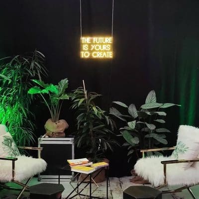Zwei weiße Stühle aus Kunstfell stehen sich in einem Raum mit Topfpflanzen, schwarzen Wänden und grüner Beleuchtung gegenüber. Auf einer Leuchtreklame darüber steht THE FUTURE IS YOURS TO CREATE. Auf einem Tisch zwischen den Stühlen liegen Bücher.