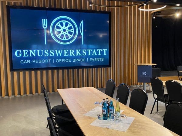 Ein moderner Besprechungsraum mit Holzwänden ist mit einem großen Bildschirm ausgestattet, auf dem Genusswerkstatt Car-Resort | Office Space | Events zu sehen ist. Ein Tisch ist mit Wasserflaschen, Gläsern und Stühlen gedeckt. Auf der rechten Seite steht ein Podium.