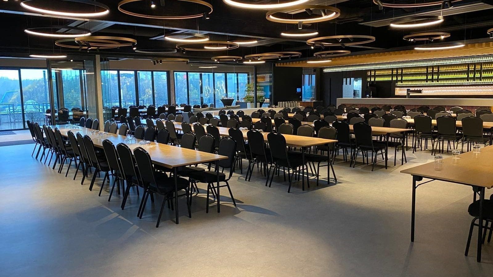 Ein geräumiger Konferenzraum mit Reihen von rechteckigen Tischen und schwarzen Stühlen, die im Stil eines Klassenzimmers angeordnet sind, großen Fenstern auf einer Seite, modernen runden Deckenleuchten und einem Tresen im hinteren Bereich.