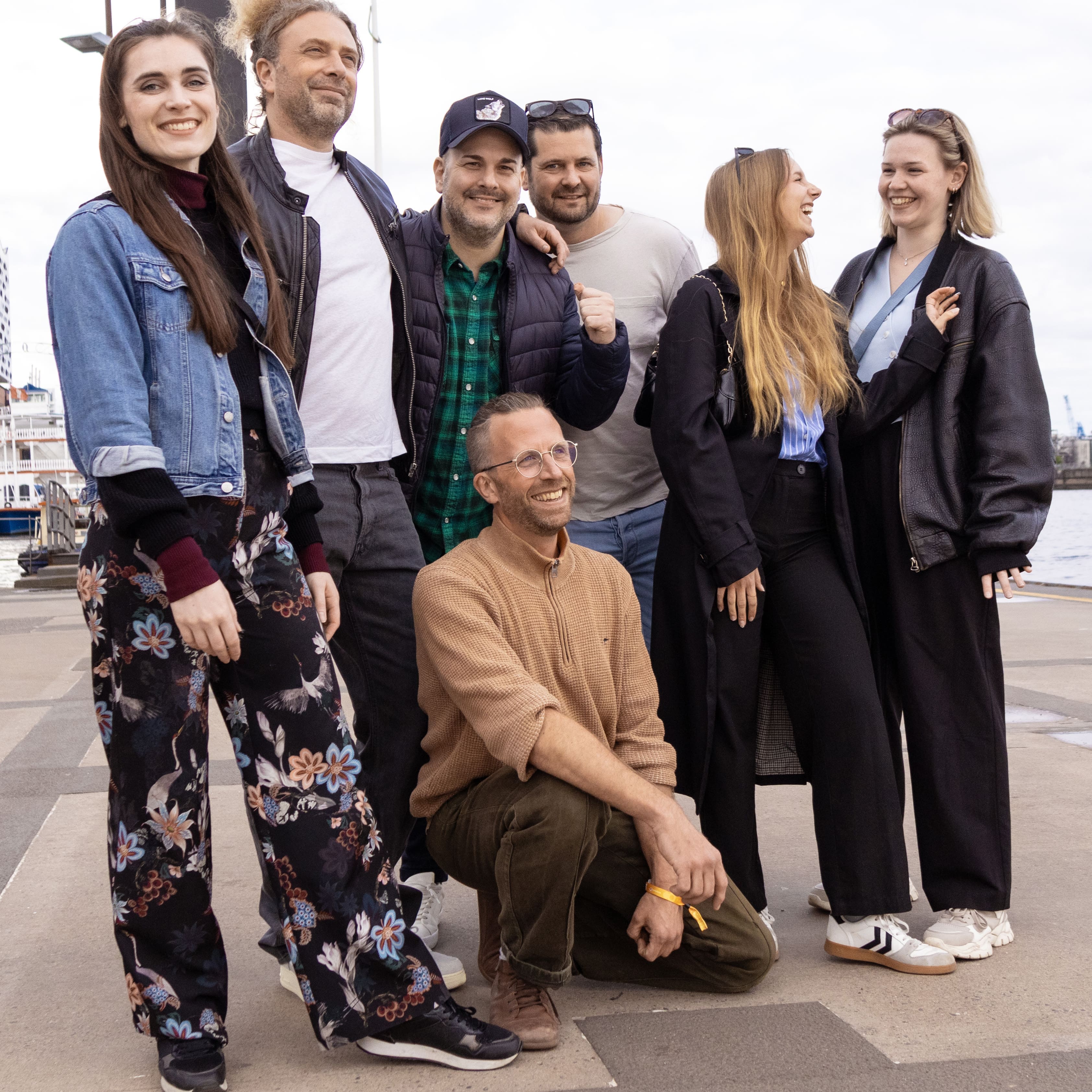 Sechs Personen posieren gemeinsam im Freien in der Nähe eines Ufers. Fünf von ihnen stehen, eine kniet vor ihnen. Die Gruppe wirkt entspannt und leger gekleidet, einige Personen lächeln und lachen. Im Hintergrund sind Boote und Wasser zu sehen.
