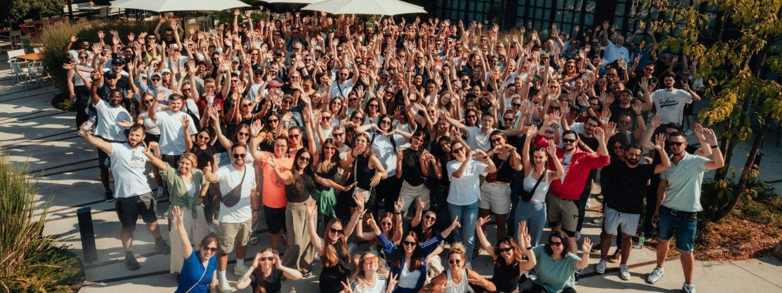 Eine große Gruppe von Menschen, die sich bei sonnigem Wetter im Freien versammelt hat, steht dicht beieinander, viele heben die Arme und lächeln, während im Hintergrund Tische, Schirme und Grünpflanzen zu sehen sind.