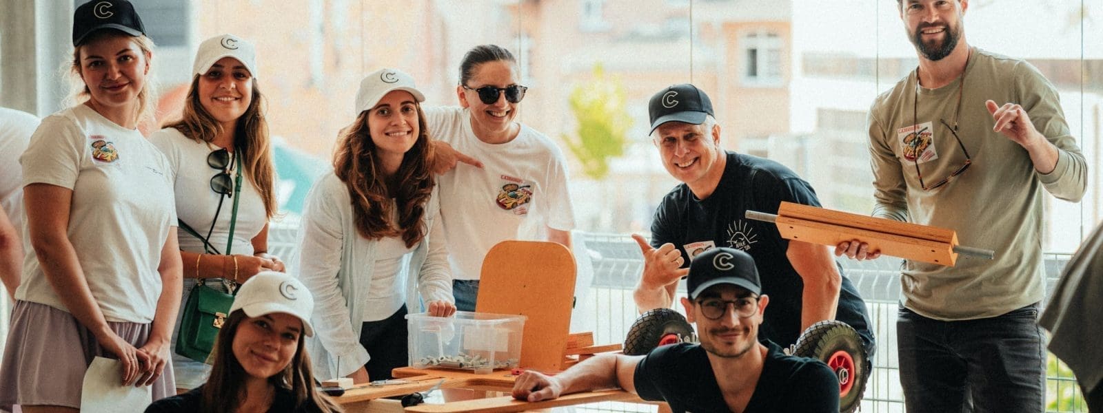 Eine Gruppe von sieben Personen, die alle weiße Mützen und Freizeitkleidung tragen, posieren in einem Innenraum um einen kleinen Tisch mit Bastelmaterial und Holzteilen. Einige sitzen, andere stehen, und sie lächeln in die Kamera.