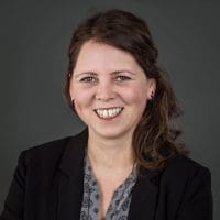 Eine Frau mit langen braunen Haaren, die einen schwarzen Blazer und eine gemusterte graue Bluse trägt, lächelt in die Kamera vor einem schlichten dunklen Hintergrund.