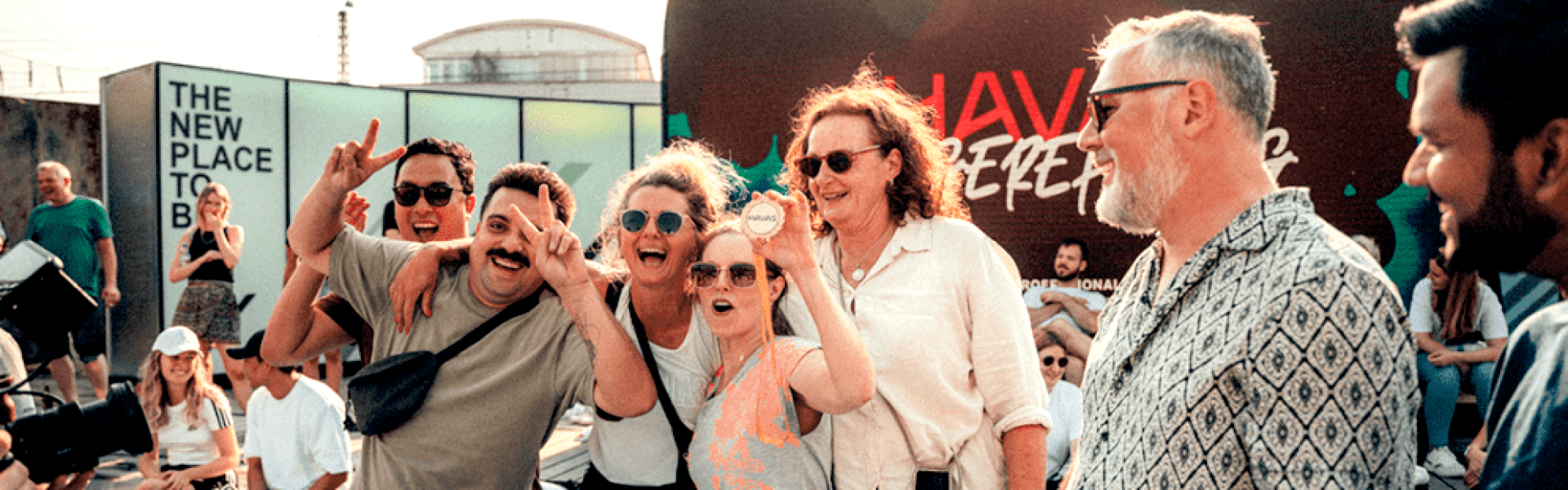 Eine Gruppe lächelnder Erwachsener posiert bei einer Veranstaltung im Freien, wobei einige Personen Handgesten machen. Andere Personen sitzen im Hintergrund, und es sind Schilder und Veranstaltungstafeln zu sehen. Die Szene wirkt lebendig und ungezwungen.