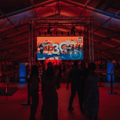 Die Menschen stehen in einem großen Veranstaltungszelt mit einer Bühne und einer großen Leinwand, auf der 