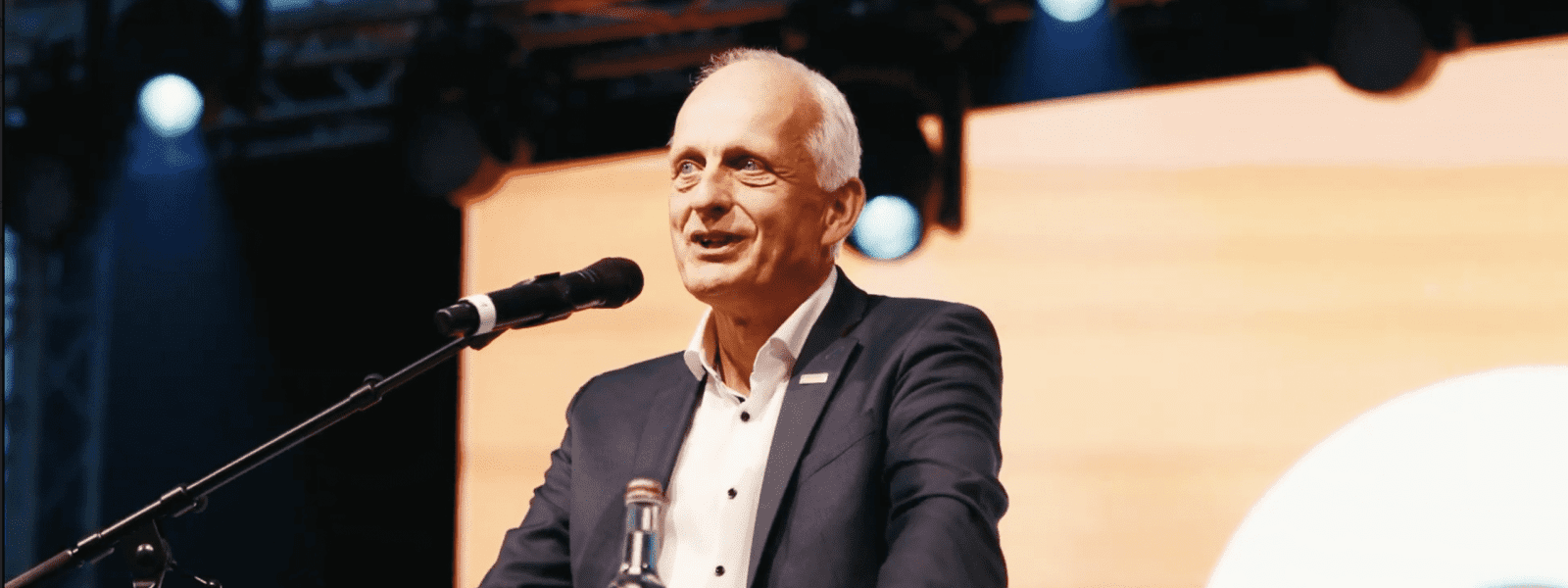 Ein Mann in einem dunklen Anzug und weißem Hemd spricht an einem Podium mit einem Mikrofon. Auf dem Podium steht eine Wasserflasche, im Hintergrund ist ein großer orangefarbener Bildschirm zu sehen, über dem die Bühnenbeleuchtung eingeschaltet ist.