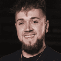 Ein junger Mann mit kurzen, gewellten braunen Haaren und einem Bart lächelt in die Kamera. Er trägt ein dunkles Hemd und eine Halskette. Hinter ihm ist ein dunkler, unscharfer Hintergrund zu sehen.