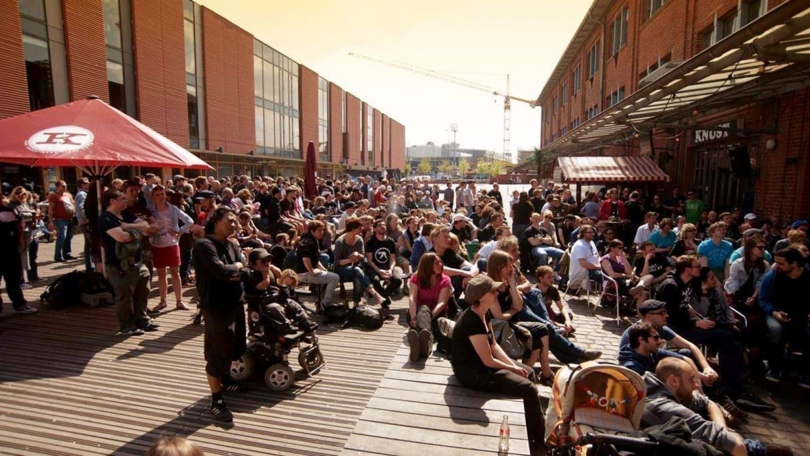 An einem sonnigen Tag sitzt und steht eine große Menschenmenge auf einem Holzdeck zwischen zwei Backsteingebäuden. Einige Leute sind mit Rollstühlen oder Kinderwagen unterwegs. Im Hintergrund sind ein roter Regenschirm und ein Baukran zu sehen.