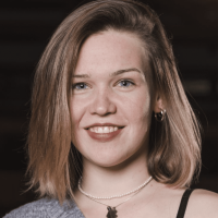 Kira Eine Frau mit schulterlangem hellbraunem Haar, die ein graues schulterfreies Oberteil, Reifohrringe, eine Perlenkette und einen schwarzen Anhänger trägt, lächelt vor einem dunklen Hintergrund in die Kamera.