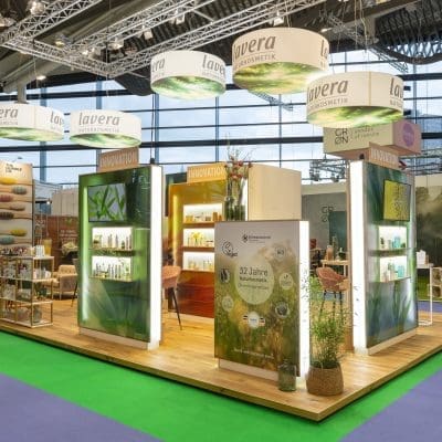Ein Messestand für Lavera-Kosmetik mit beleuchteten Displays mit Produkten, Werbebannern, Regalen mit verschiedenen Artikeln und einem modernen Design in Holzoptik in einer geräumigen Ausstellungshalle.