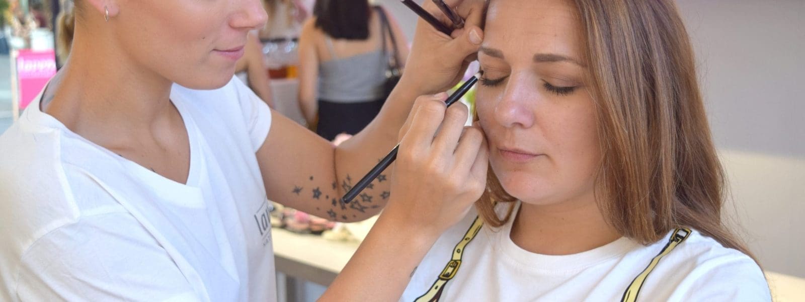 Eine Visagistin trägt Lidschatten auf eine sitzende Frau auf, die ihre Augen geschlossen hat. Der Schauplatz scheint ein Schönheitssalon mit Menschen und einer Leuchtreklame im Hintergrund zu sein.