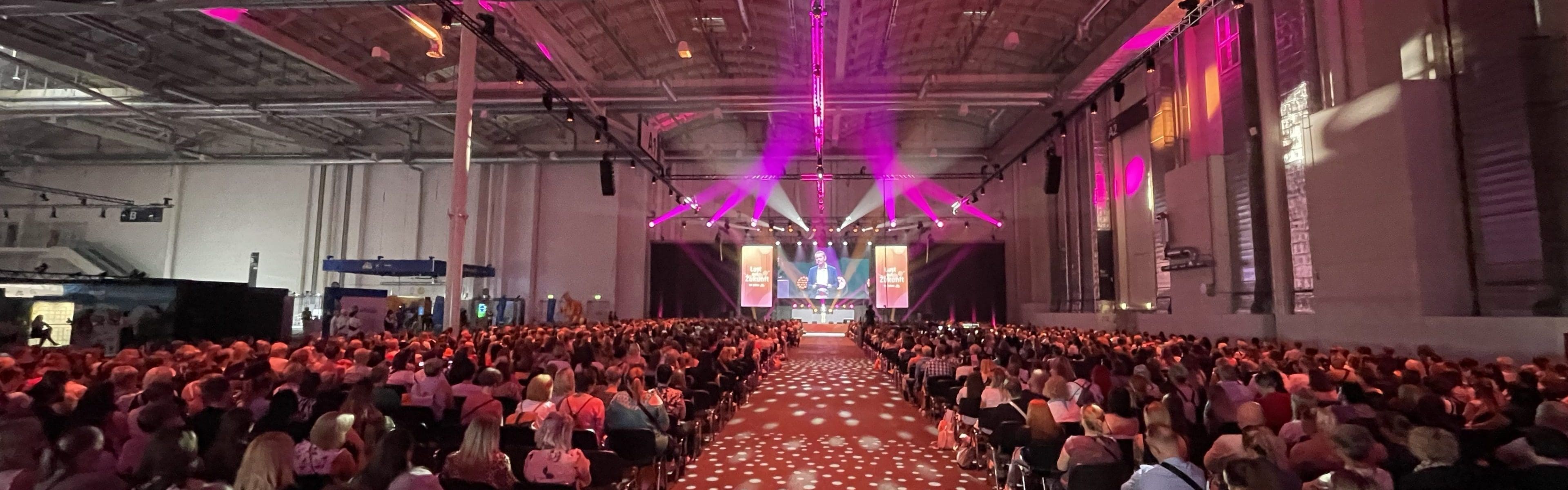 Ein großes Publikum sitzt in Reihen vor einer hell erleuchteten Bühne im vorderen Teil einer geräumigen Veranstaltungshalle. Rosa und weiße Lichter erzeugen Muster an der Decke und auf dem Boden. Auf beiden Seiten der Bühne hängen Transparente.
