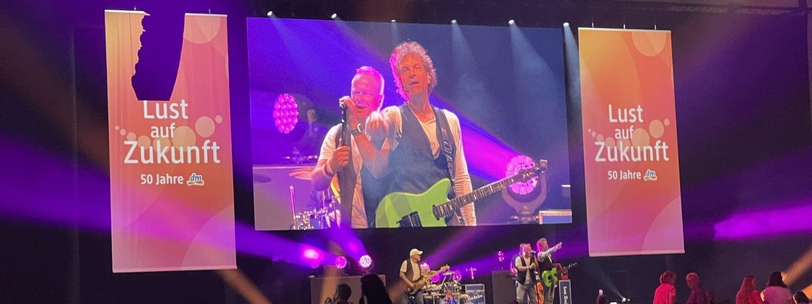 Eine Band spielt auf einer Bühne mit rosa und lila Beleuchtung. Große Bildschirme zeigen die Musiker und den Text Lust auf Zukunft 50 Jahre. Reihen von leeren rosa Stühlen füllen den Vordergrund der Halle.