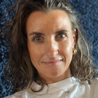 MArlen Lange – Porsche Eine Frau mit langen braunen Haaren und hellen Augen trägt ein weißes Oberteil und goldene Ohrringe und blickt direkt in die Kamera. Der Hintergrund ist dunkelblau und hat ein strukturiertes Aussehen.