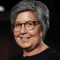 Eine Person mittleren Alters mit kurzen grauen Haaren, die eine Brille und ein dunkles Oberteil trägt, lächelt in die Kamera vor einem dunklen Hintergrund.