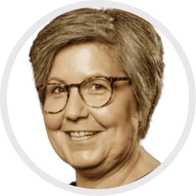 Eine Frau mit kurzen grauen Haaren und Brille lächelt in die Kamera. Sie trägt ein helles Oberteil und erscheint in einem kreisförmigen Ausschnitt vor einem schlichten weißen Hintergrund.
