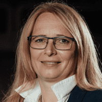 Eine Frau mit langen blonden Haaren und Brille ist in einer Nahaufnahme zu sehen. Sie trägt ein weißes Hemd mit Kragen und eine dunkle Jacke, und sie lächelt leicht. Der Hintergrund ist dunkel und unscharf.