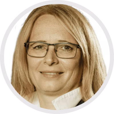 Eine Frau mit langem blondem Haar und Brille ist der Kamera zugewandt und trägt ein weißes Hemd mit Kragen und eine dunkle Jacke. Sie lächelt leicht. Das Bild wird von einem weißen, kreisförmigen Rahmen umrahmt.