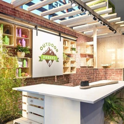 Ein Stand mit einer weißen Holztheke, Backsteinwänden und Holzregalen stellt bunte Kerzen aus. Auf einem großen Schild steht Outdoor Freakz mit einer Berggrafik. Es gibt Pflanzen und natürliches Licht fällt von oben ein.
