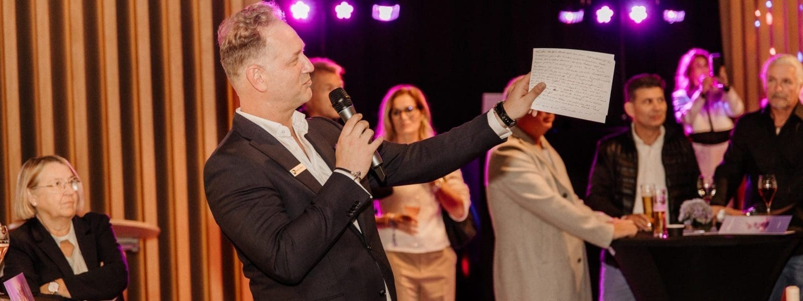 Ein Mann im Anzug spricht in ein Mikrofon und hält ein Blatt Papier in der Hand, während er vor einer Gruppe von Menschen bei einer Indoor-Veranstaltung steht. Mehrere Teilnehmer schauen zu, während auf den Cocktailtischen in der Nähe Getränke und Snacks stehen.