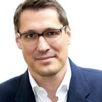 Ein Mann mit kurzen braunen Haaren und Brille, der eine dunkle Anzugsjacke und ein weißes Hemd mit Kragen trägt und vor einem schlichten weißen Hintergrund direkt in die Kamera schaut.