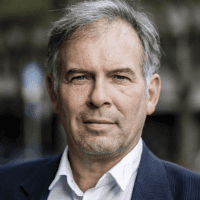 Ein Mann mittleren Alters mit grauem Haar und hellem Bart trägt eine dunkle Anzugsjacke und ein weißes Hemd. Er steht im Freien und hat einen neutralen Gesichtsausdruck, während die Bäume im Hintergrund verschwimmen.