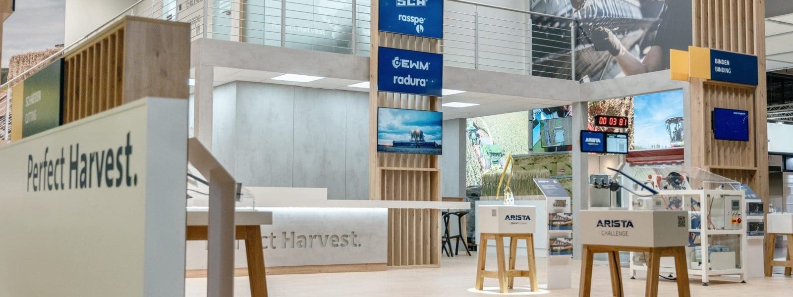 Messestand mit Holzakzenten, hoher Decke, Ausstellungstischen, Markenschildern und Bildern von Maschinen. Erhöhte Plattform mit Glasgeländer und Treppe im Hintergrund. Der Boden ist aus hellem Holz.