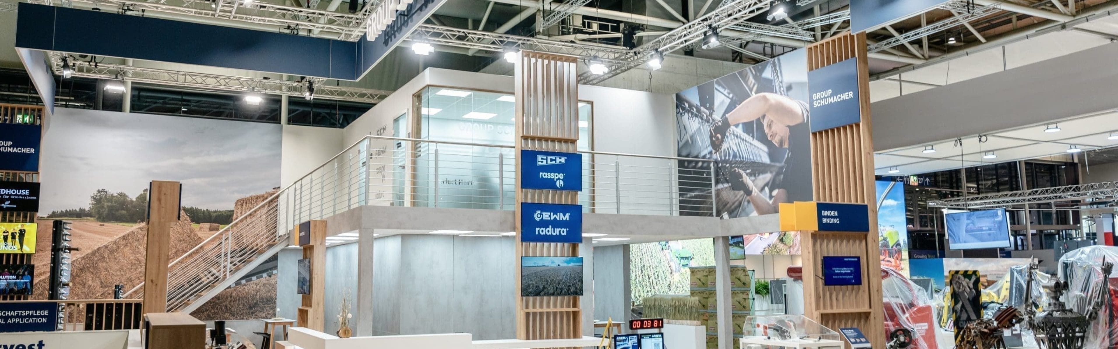 Ein moderner Messestand für die Schumacher-Gruppe zeichnet sich durch Holz- und Metallakzente, digitale Bildschirme, landwirtschaftliche Displays und eine zweite Ebene mit einem Glasgeländer aus. Schilder und Maschinen heben landwirtschaftliche Technik und Ausrüstung hervor.