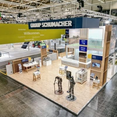 Ein großer, moderner Messestand für die Schumacher-Gruppe mit interaktiven Displays, Informationsschildern, Sitzgelegenheiten, Produktproben und lebensgroßen Metallskulpturen auf einem Boden in Holzoptik in einer Ausstellungshalle.