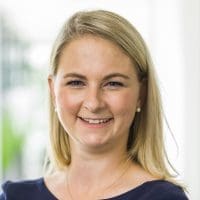 Eine Frau mit glattem blondem Haar, die ein marineblaues Oberteil und Perlenohrringe trägt, lächelt in einer hell erleuchteten Innenumgebung in die Kamera.