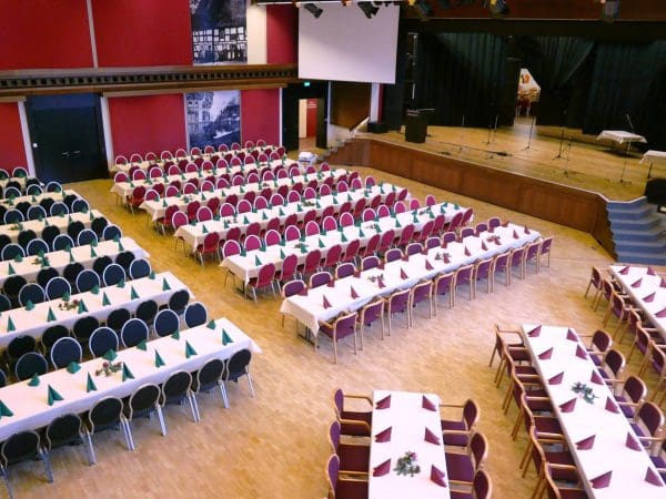 Großer Veranstaltungssaal mit Reihen von rechteckigen Tischen mit weißen Tischdecken und grünen Servietten, umgeben von roten und schwarzen Stühlen. An der Stirnseite befindet sich eine Bühne mit Mikrofonen und einer Treppe, die zu ihr hinaufführt.