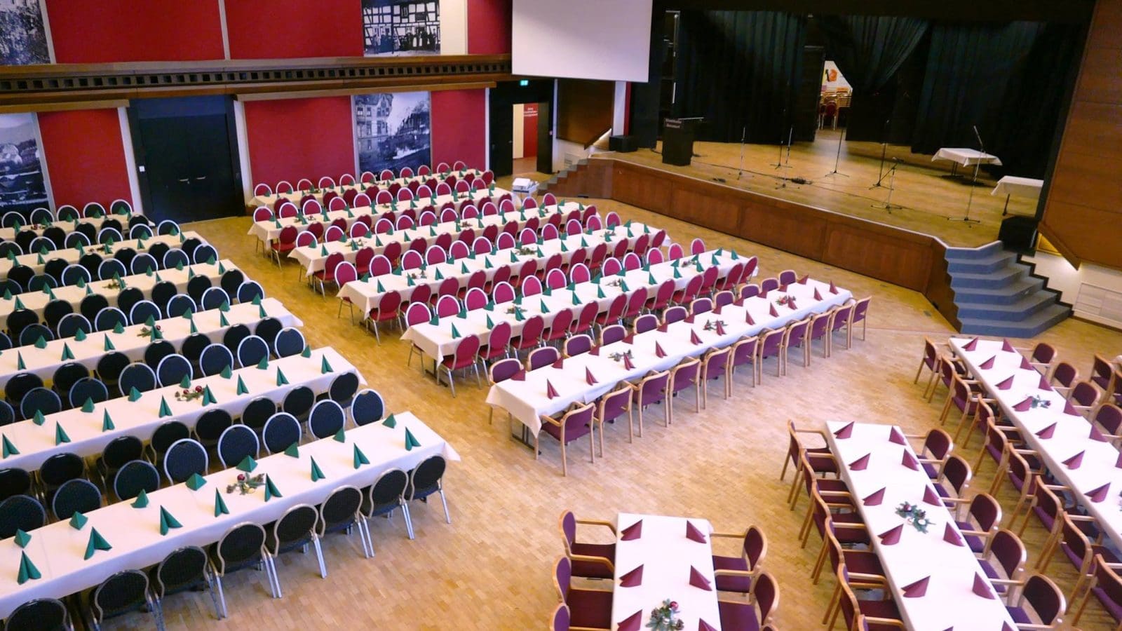 Ein großer Veranstaltungssaal mit Reihen von rechteckigen Tischen mit weißen Tischdecken, grünen Servietten und Blumenschmuck, vielen roten Stühlen, einer Bühne mit Mikrofonen und holzgetäfelten Böden und Wänden.