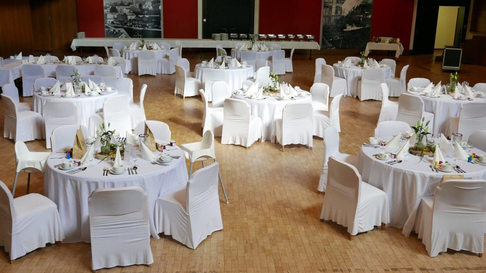 Ein großer Bankettsaal mit runden Tischen mit weißen Tischdecken und Stühlen, die für eine formelle Veranstaltung gedeckt sind. Jeder Tisch hat gefaltete Servietten, Besteck, Gläser und einfache Tafelaufsätze. Der Boden ist aus Holz.