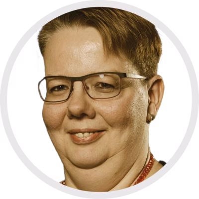 Eine Person mit kurzen braunen Haaren, einer Brille und einem Nasenpiercing blickt in die Kamera und lächelt leicht. Der Hintergrund ist einfarbig weiß, und das Bild wird von einem kreisförmigen Rahmen eingerahmt.