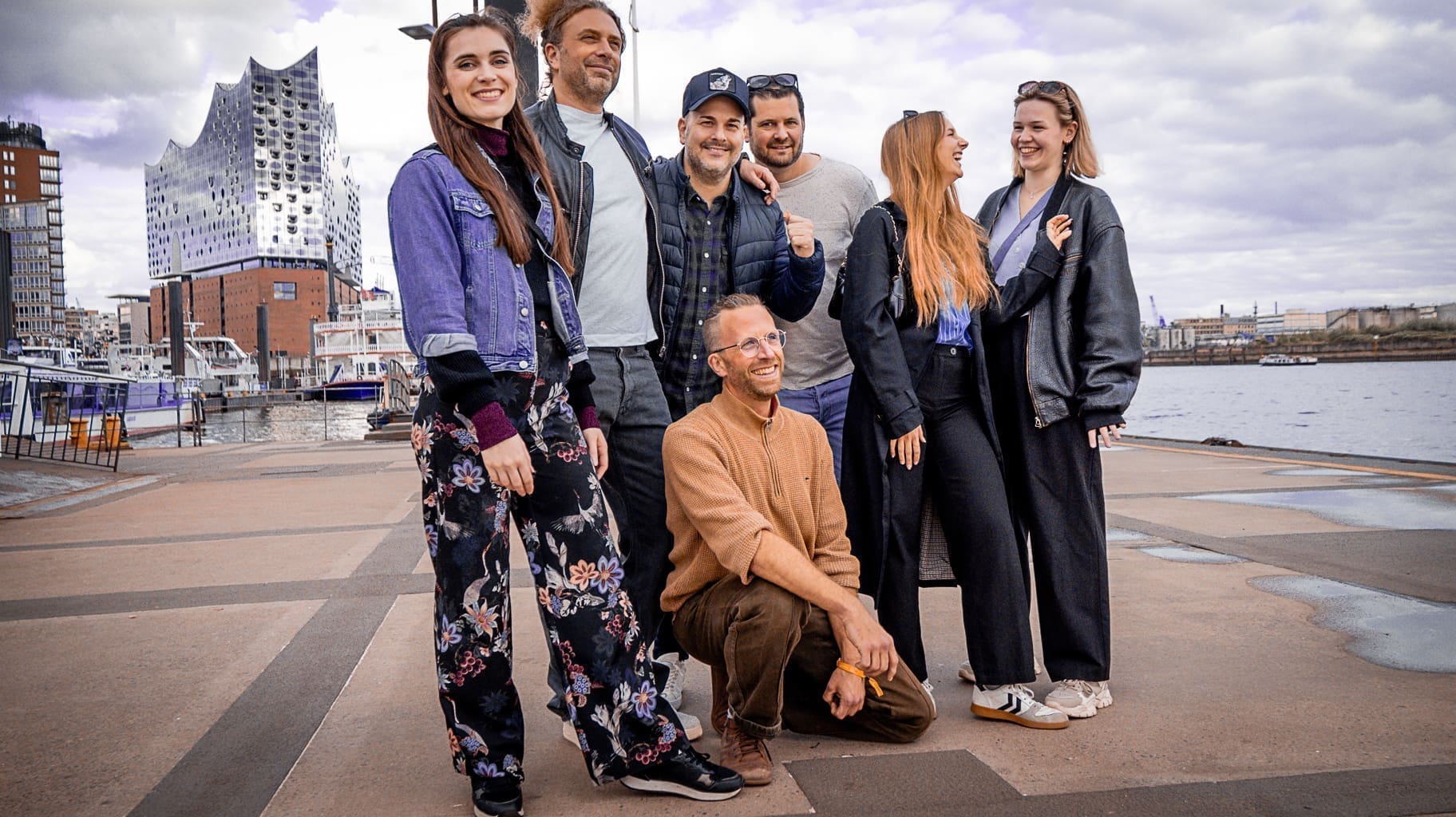 Eine Gruppe von sieben Personen posiert und lächelt im Freien in der Nähe einer Uferpromenade. Im Hintergrund sind moderne Gebäude zu sehen, darunter die Elbphilharmonie in Hamburg, und der Himmel ist leicht bewölkt.