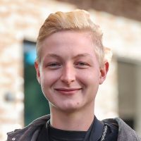 Tim Eine Person mit kurzem, blondem Haar lächelt in die Kamera. Sie trägt ein dunkles Oberteil und steht im Freien vor einem unscharfen Hintergrund mit einem Gebäude und Fenstern.