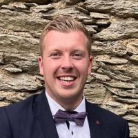 Tim Blöcher Ein Mann in dunkler Anzugsjacke, weißem Hemd und gemusterter Fliege steht lächelnd vor einer Steinmauer.