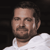 Ein Mann mit kurzen braunen Haaren und gestutztem Bart trägt einen weißen Kapuzenpullover. Er ist der Kamera zugewandt und hat einen neutralen Gesichtsausdruck. Der Hintergrund ist dunkel und unscharf.