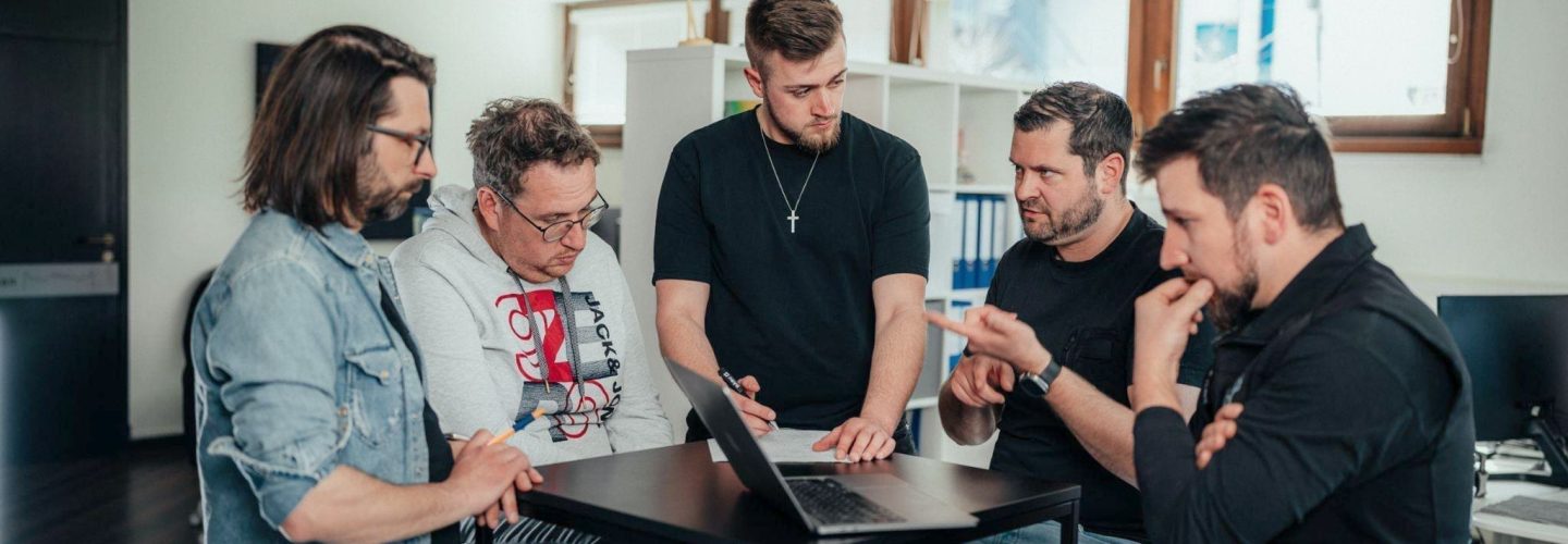 Fünf Männer sitzen und stehen um einen Tisch herum, schauen gemeinsam auf einen Laptop-Bildschirm und scheinen in einer modernen Büroumgebung etwas zu besprechen oder zu überprüfen.