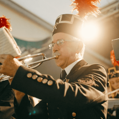Ein älterer Mann in einer geschmückten Uniform und einem Hut mit roter Feder spielt ein Blechblasinstrument in einer Kapelle, während im Hintergrund andere Musiker in ähnlicher Kleidung zu sehen sind. Hinter ihm scheint das Sonnenlicht.