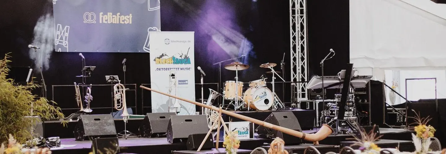 Ein dekoriertes Veranstaltungszelt mit einer Bühne, die mit Musikinstrumenten, Lautsprechern und Beleuchtung ausgestattet ist. Die Tische sind mit Tellern, Tassen und Blumenarrangements sowie großen Brezeln auf Ständern für jeden Tisch gedeckt.