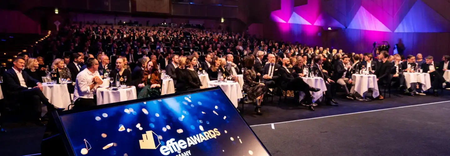 event-marketing-agentur-duisburg Ein großes, formell gekleidetes Publikum sitzt an runden Tischen in einem schwach beleuchteten Veranstaltungssaal. Im Vordergrund zeigt ein Bildschirm die Effie Awards Germany mit konfettiartigen Grafiken.