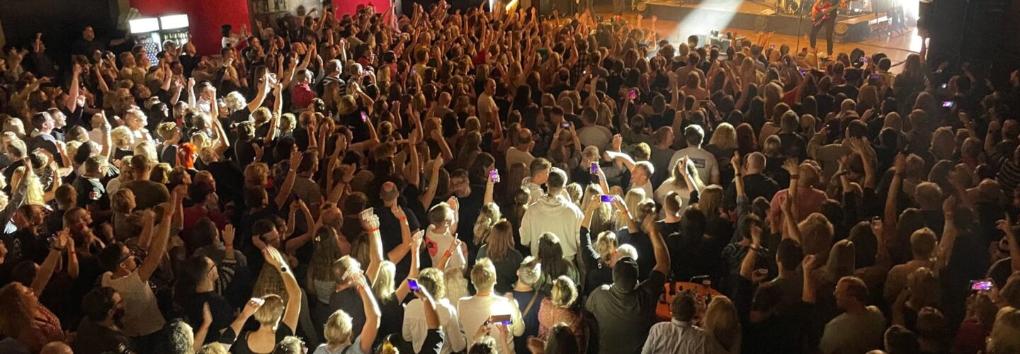 Eine große Menschenmenge füllt einen überdachten Konzertsaal und blickt auf eine gut beleuchtete Bühne, auf der eine Band auftritt. Die Zuhörer stehen dicht beieinander, viele mit erhobenen Armen, während das Bühnenlicht in die Menge scheint.