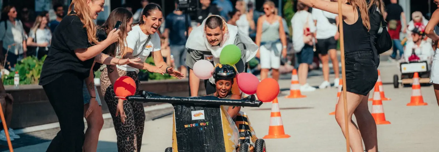 event-marketing-agentur-greifswald Eine Gruppe von Menschen jubelt einer Person zu, die einen Helm trägt und in einem kleinen, selbst gebauten und mit Luftballons geschmückten Wagen fährt. Orangefarbene Verkehrskegel säumen den Weg, und im Hintergrund schaut eine Menschenmenge zu.