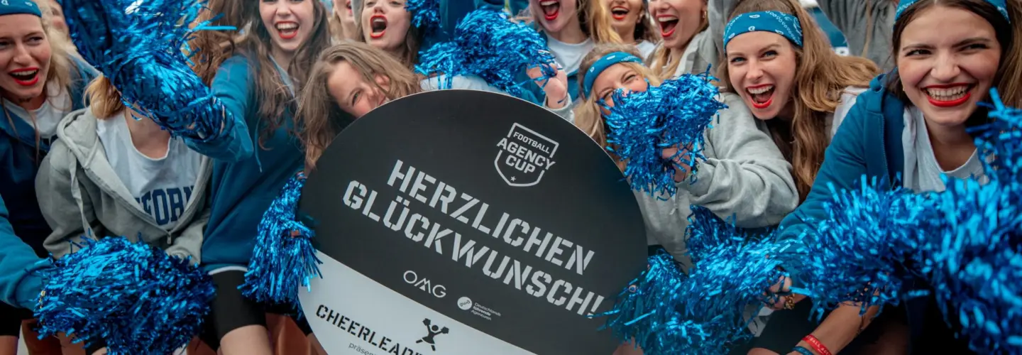 Eine Gruppe von Cheerleadern in blauen Uniformen und Stirnbändern lächelt und hebt blaue Pom-Poms hoch, während sie ein rundes Schild mit der Aufschrift "Herzlichen Glückwunsch! Cheerleader Cup 2023".