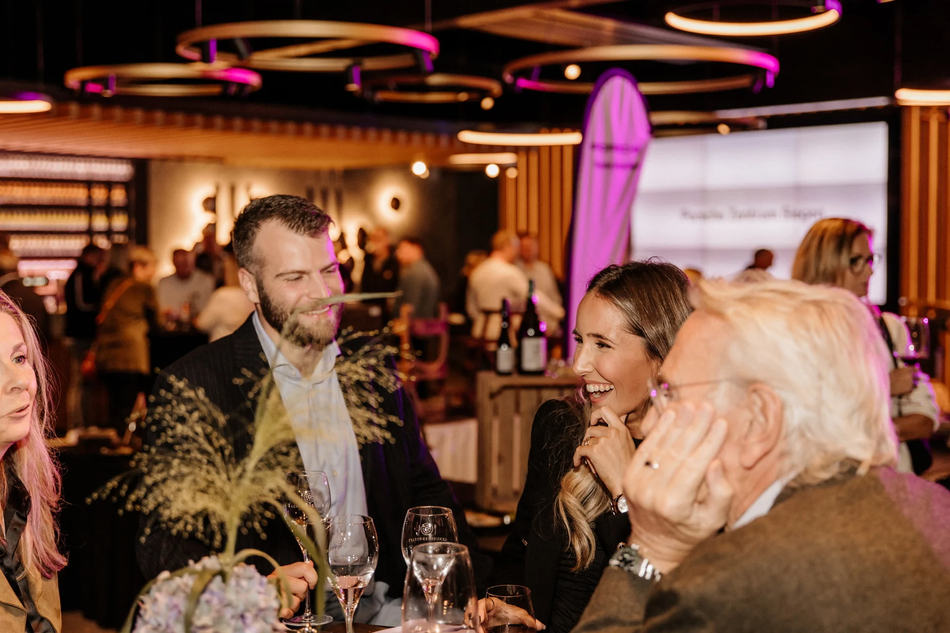 event-marketing-agentur-lueneburg Eine Gruppe von Menschen in halb-formeller Kleidung steht um einen Tisch mit Weingläsern bei einer Indoor-Veranstaltung und unterhält sich. Der Hintergrund ist mit violettem Licht warm beleuchtet und andere Gäste sind zu sehen.