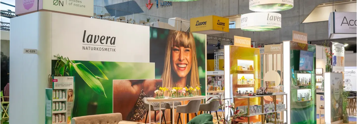 Ein Lavera Naturkosmetik-Messestand verfügt über Produktregale, eine Sitzecke mit grünen Stühlen und Sofas, einen Tisch mit Blumen, Markenschilder und große Bilder der Natur und einer lächelnden Frau. Deckenleuchten und Banner hängen über dem Stand.