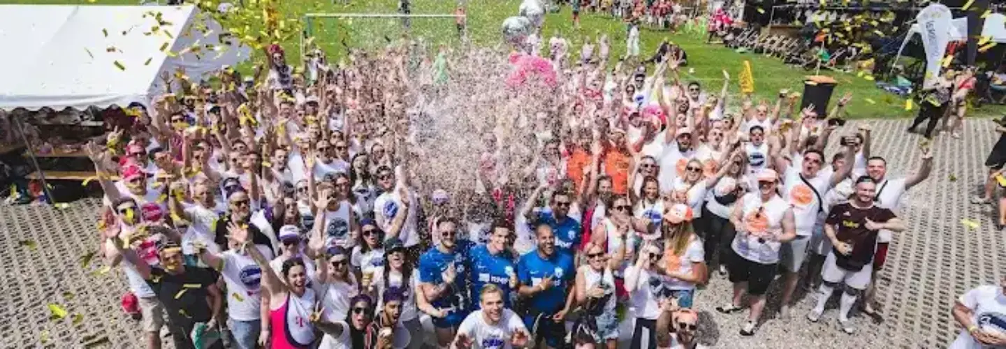 event-marketing-agentur-schwerin Eine große Gruppe von Menschen steht im Freien, viele tragen passende T-Shirts, feiern mit Konfetti und verspritzen Getränke. Die Veranstaltung findet an einem sonnigen Tag statt, mit Zelten und Sportplätzen im Hintergrund.