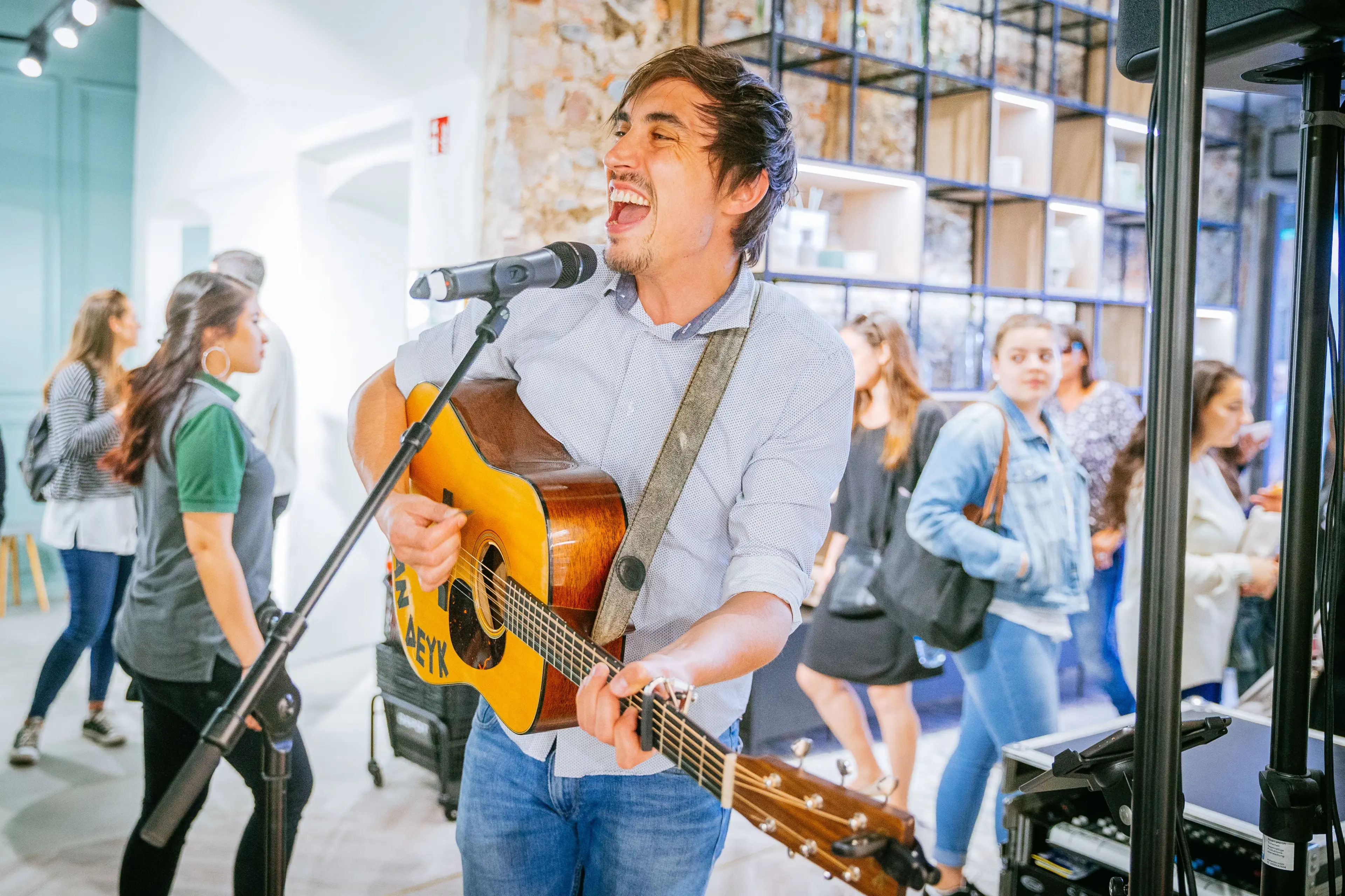 Ein Mann spielt eine akustische Gitarre und singt in ein Mikrofon in einem hellen Innenraum. Mehrere Menschen stehen und gehen im Hintergrund herum, einige schauen zu, andere unterhalten sich.