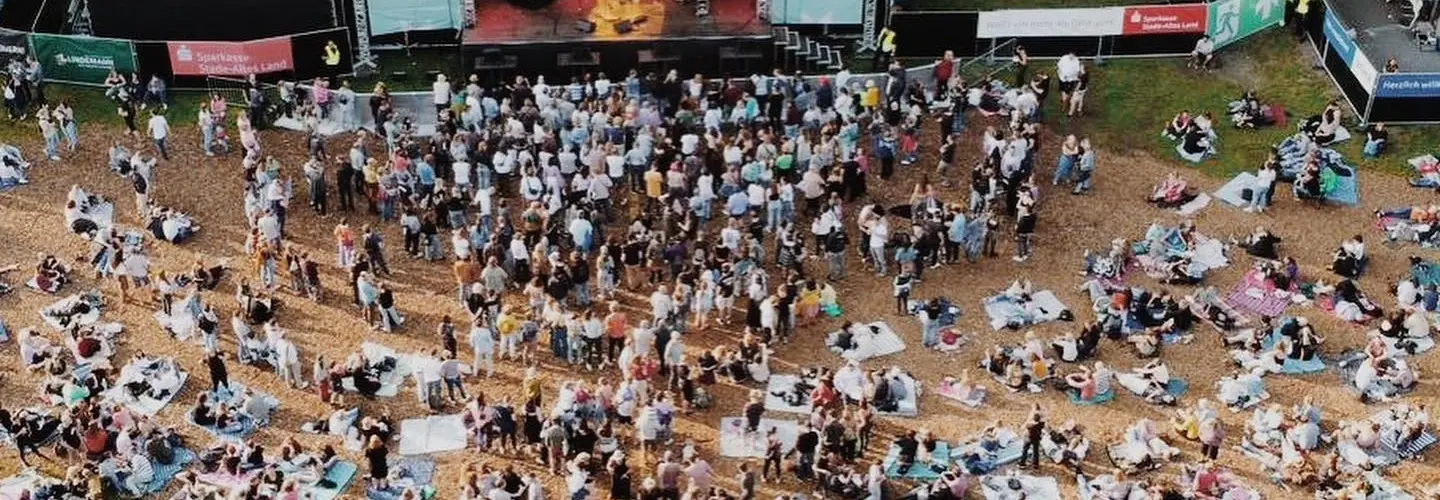 Luftaufnahme eines Konzerts im Freien mit einer Bühne, Künstlern und einem Publikum, das auf Decken auf einer offenen, von Bäumen umgebenen Wiese steht und sitzt. Die Menschen sind vor der Bühne versammelt, einige sitzen weiter hinten.